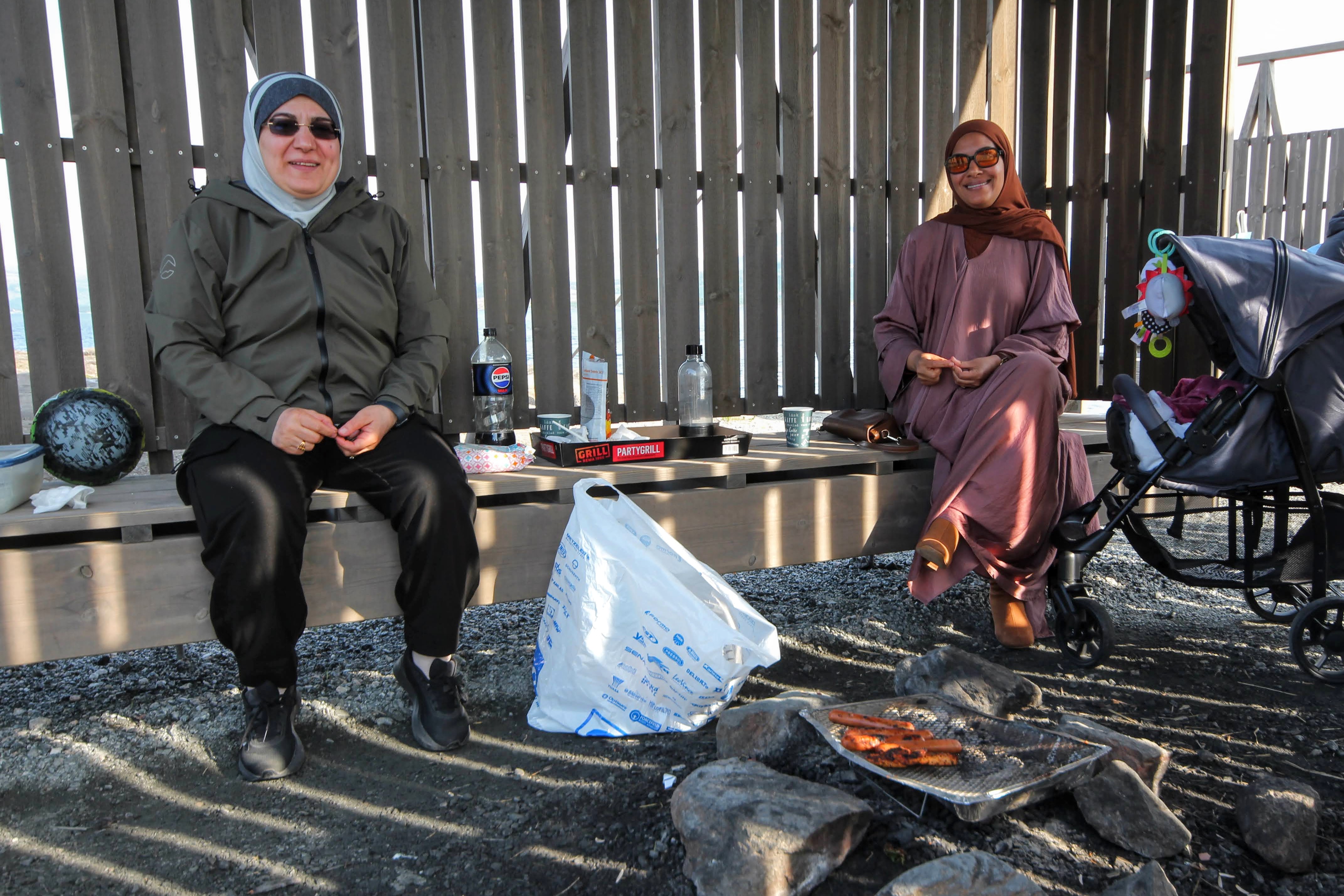 To damer sitter på en benk. Det står en barnevogn ved siden av. Foran står en engangsgrill med grillmat.